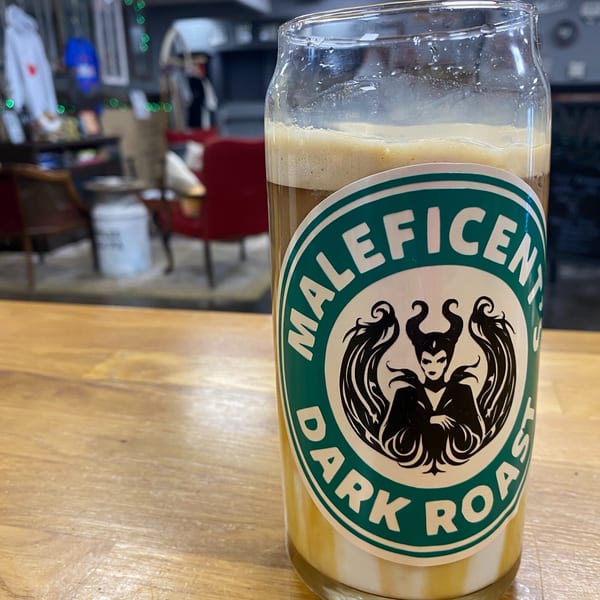 A glass of layered coffee drink with a "Maleficent Dark Roast" sticker on a wooden table.