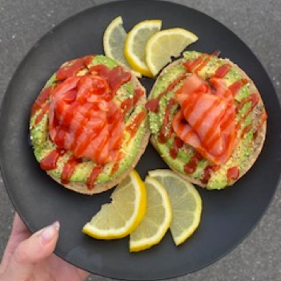 Two avocado toasts with smoked salmon, sriracha, and lemon wedges on a black plate.