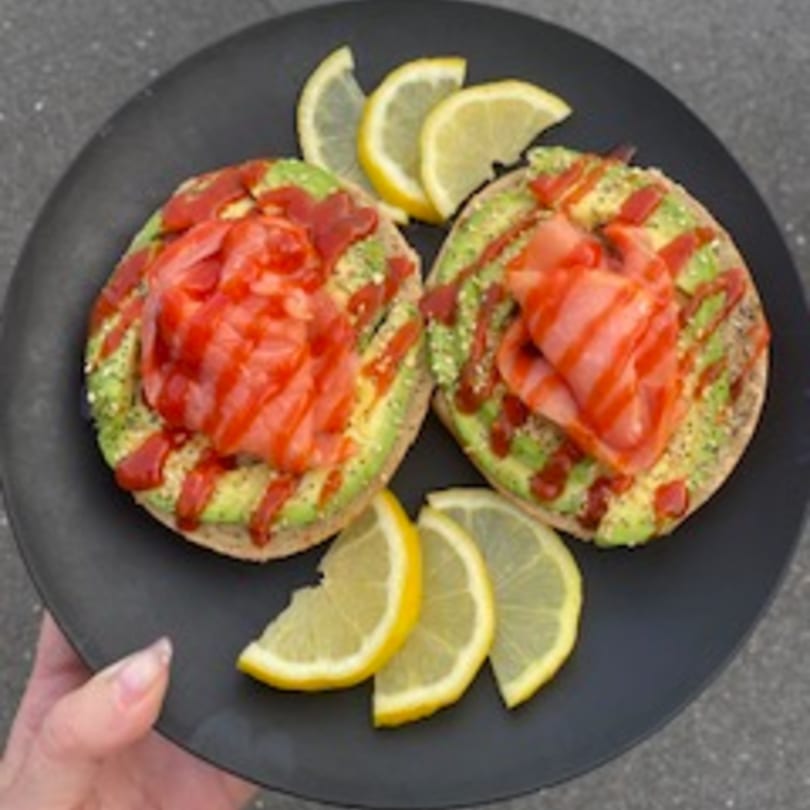 Two avocado toasts with smoked salmon, sriracha, and lemon wedges on a black plate.