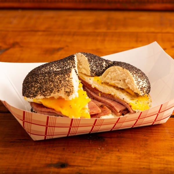 Poppy seed bagel with egg, cheese, and sliced meat in a paper tray on a wooden table.