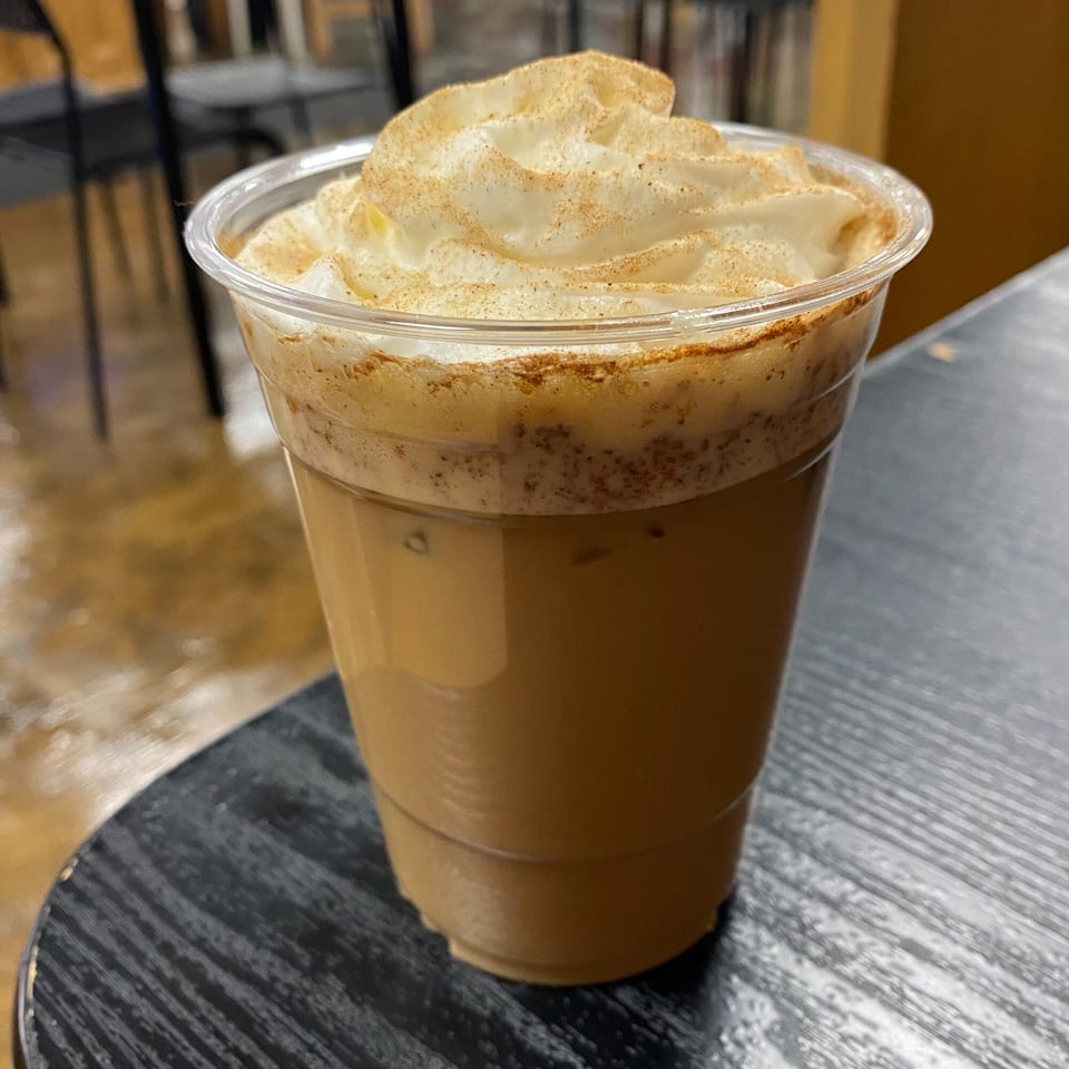 Iced coffee with whipped cream and cinnamon sprinkles in a clear plastic cup on a dark table.
