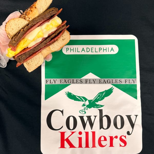 A Redhouse Bagels sandwich with egg, cheese, and meat, held above a "Cowboy Killers" sign.