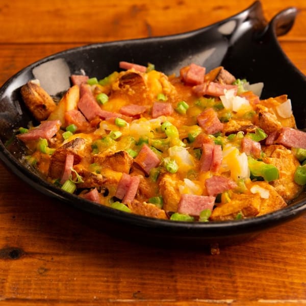 A black bowl of loaded breakfast potatoes with melted cheese, diced ham, and green onions.