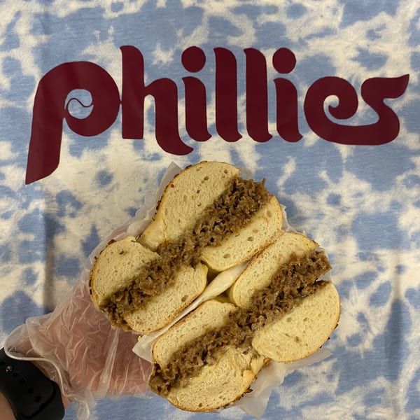 A Redhouse Bagels cheesesteak bagel, cut in half, held by a gloved hand over a Phillies shirt.