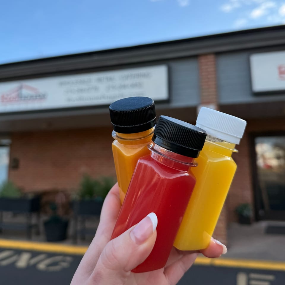 A hand holds three small bottles of colorful juice, red and yellow, in front of Redhouse Bagels.